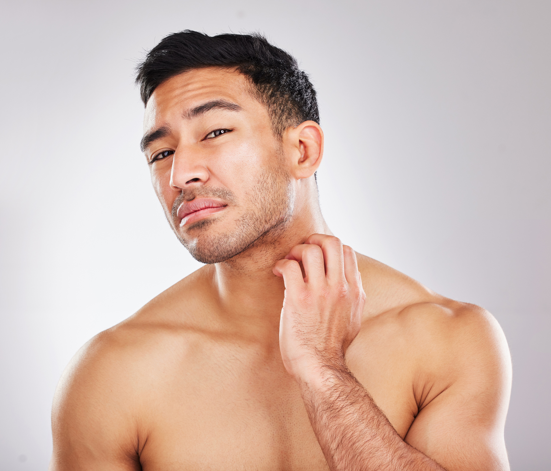 Portrait of a young man checking his skin and looking unhappy during his skincare routine against a grey background